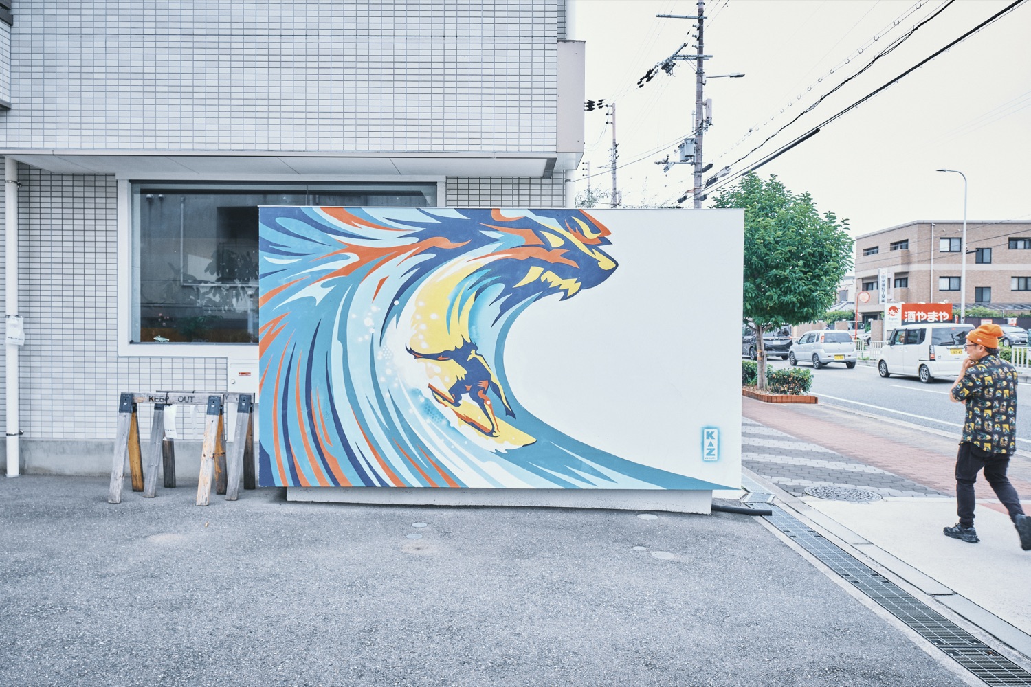 建物の壁面に描かれた、波に乗るサーファーのミューラルアートと、それを見るカズ・オオモリ